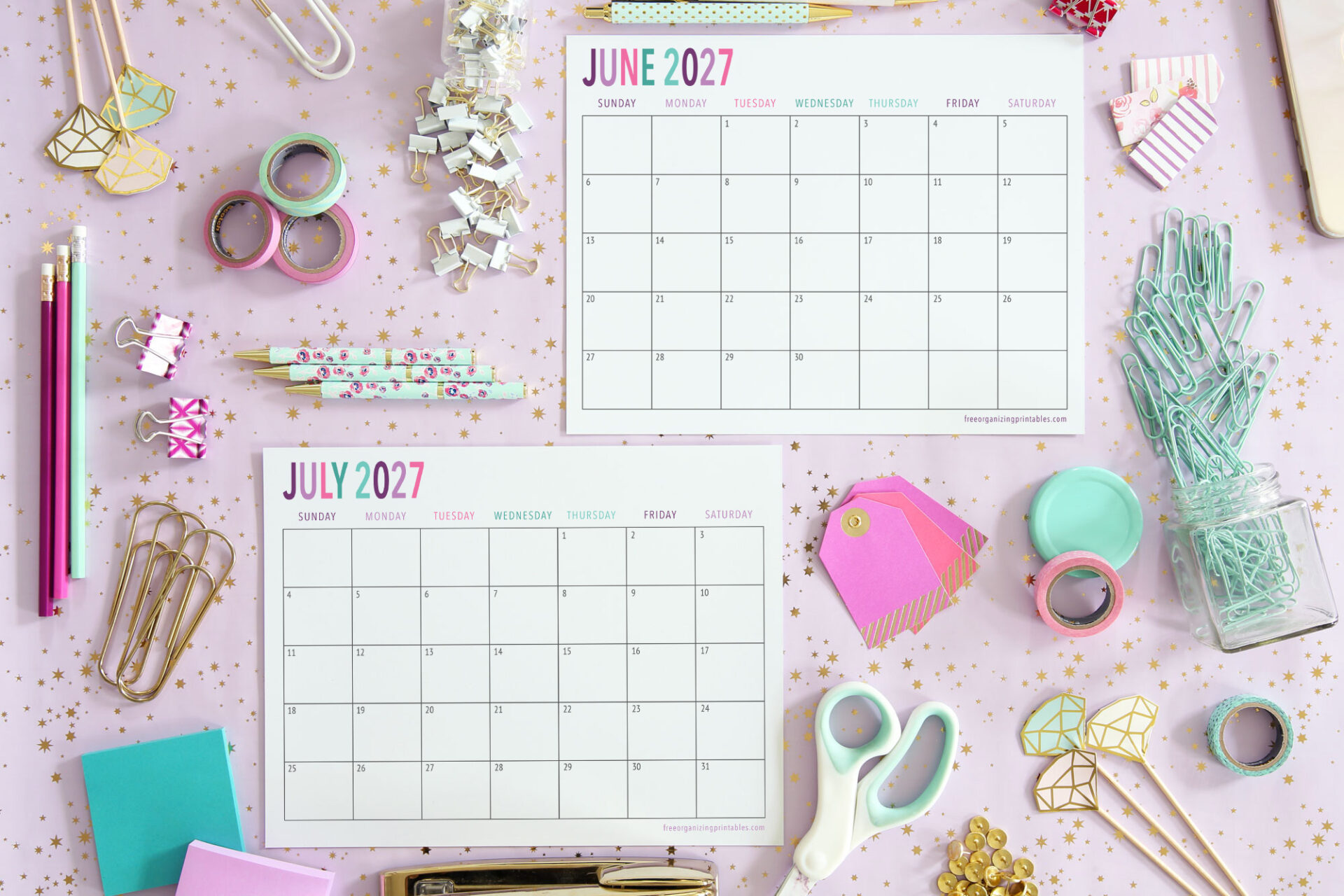 Two monthly calendars for June and July 2027 on a pink desk with gold stars, surrounded by colorful stationery like pens, clips, washi tape, scissors, and sticky notes.