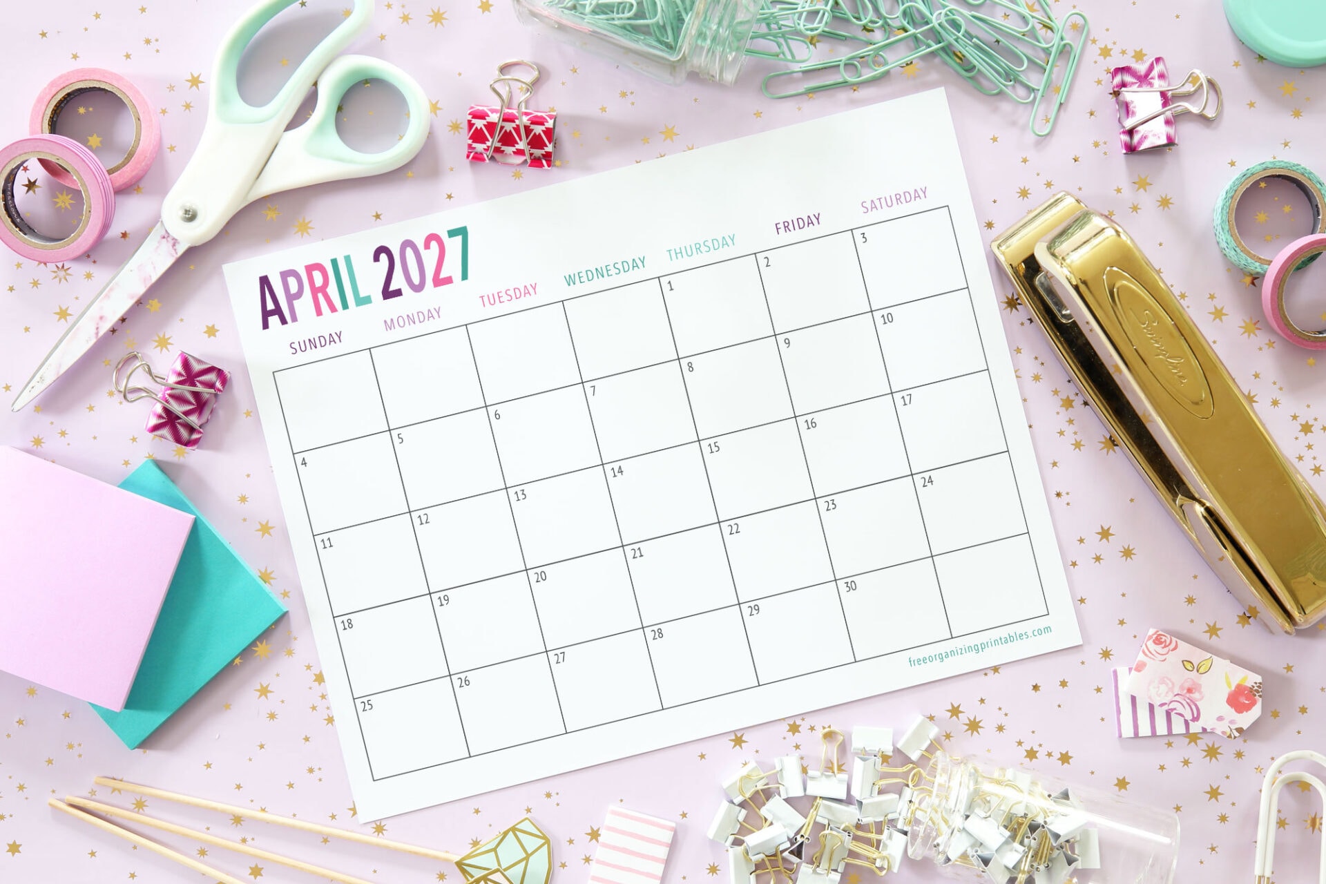 A blank April 2027 calendar lies on a pastel desk with scissors, stapler, tape, stamps, colorful paper, and office supplies scattered around.