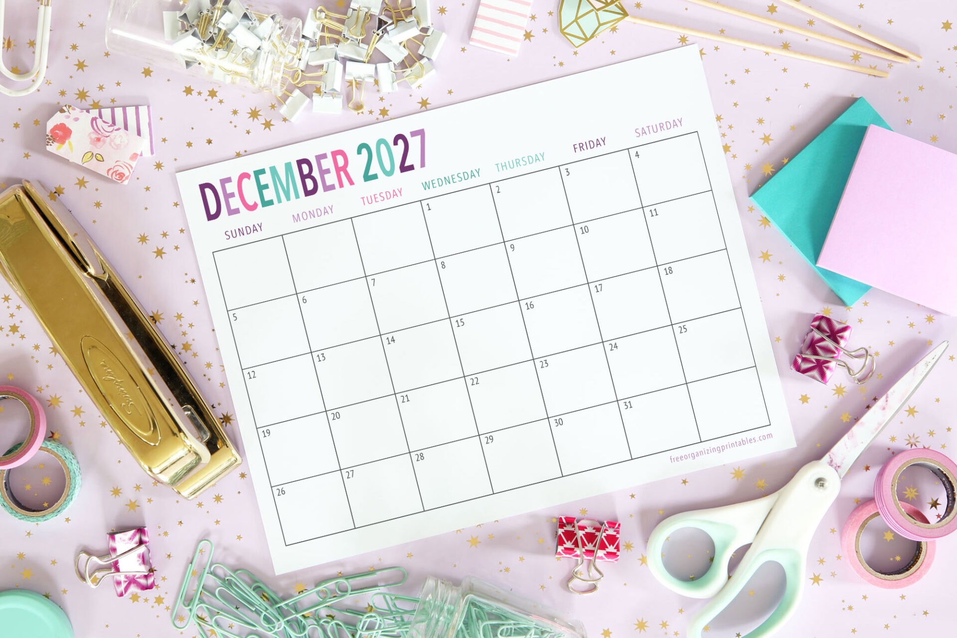 A December 2027 calendar is surrounded by office supplies, including a stapler, scissors, tape, binder clips, paper clips, sticky notes, and decorative items on a white surface with pink and gold stars.