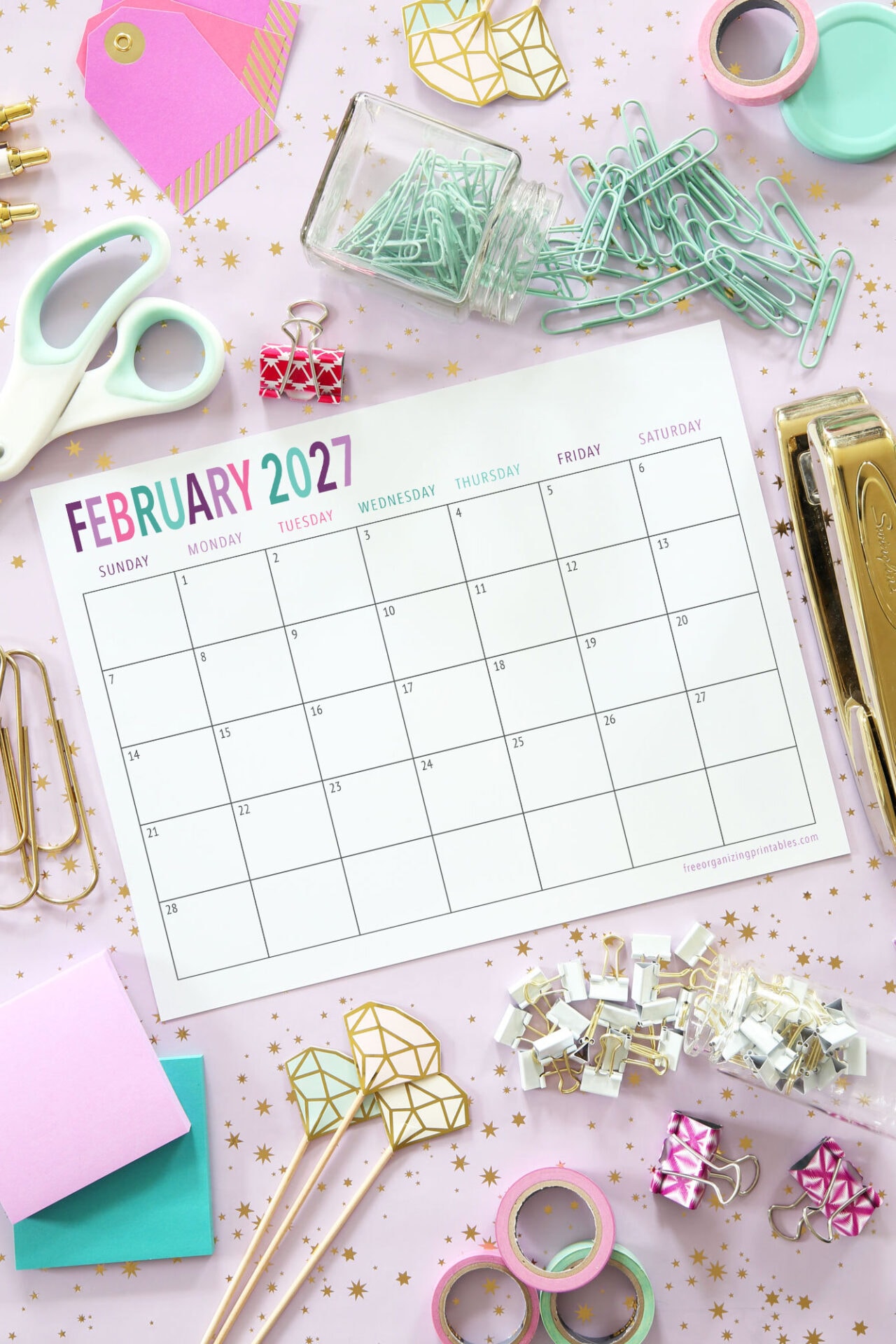 A colorful flat lay of a blank February 2027 calendar surrounded by office supplies like scissors, paper clips, binder clips, tape, sticky notes, and a stapler on a star-patterned background.
