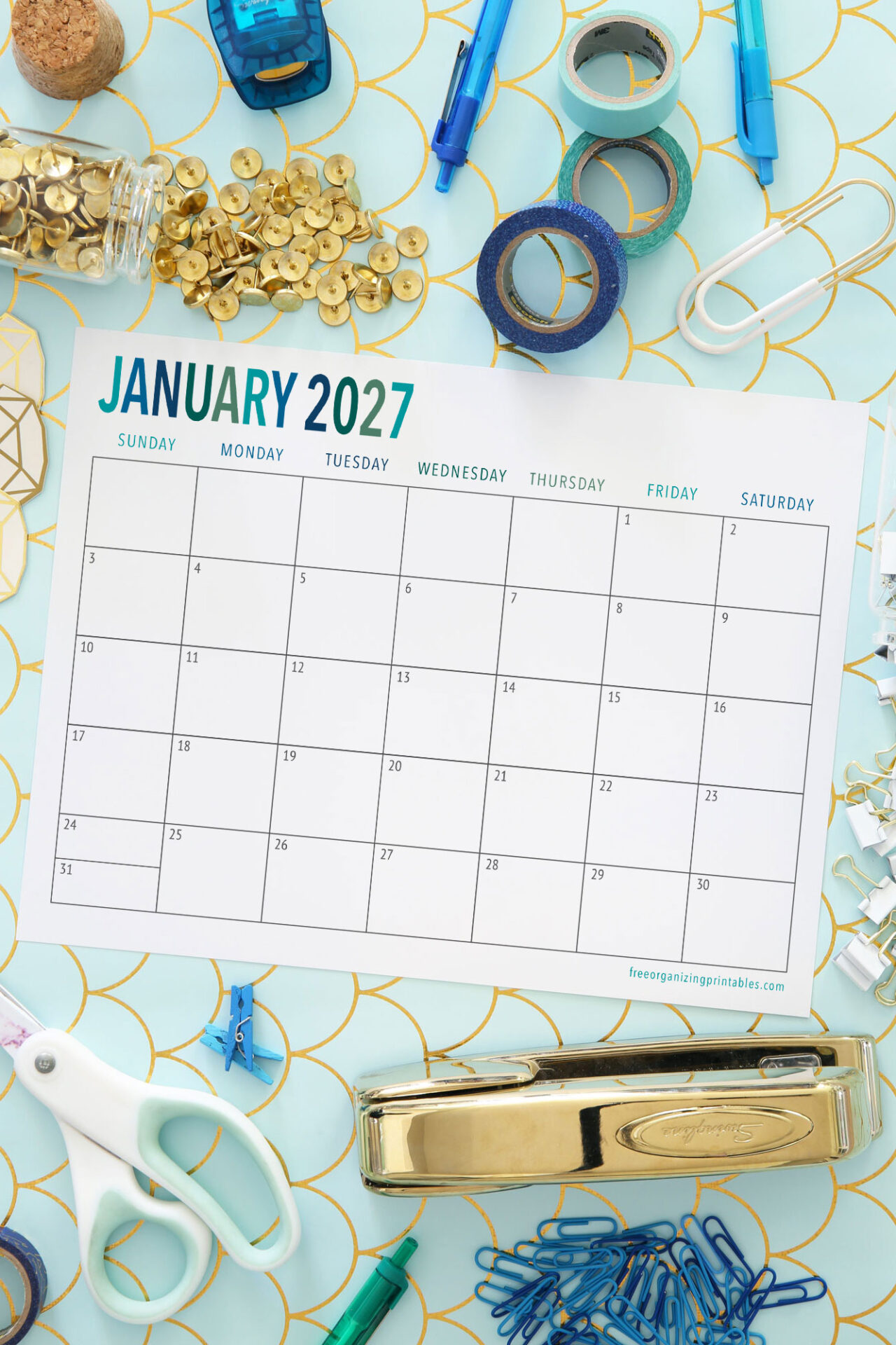 A blank January 2027 calendar is surrounded by office supplies like scissors, a stapler, paperclips, push pins, binder clips, and rolls of tape on a blue and gold patterned background.