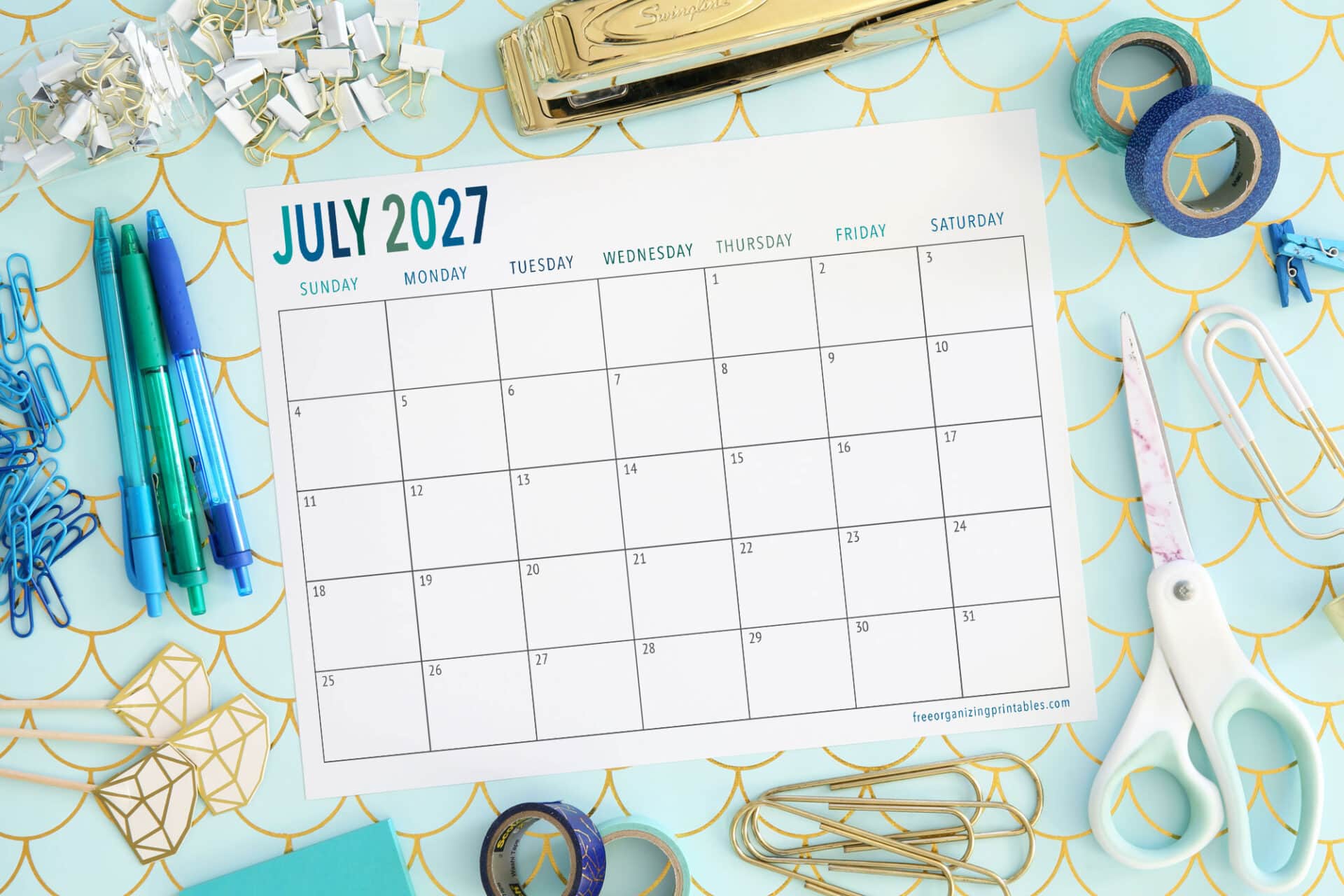 A July 2027 calendar is surrounded by office supplies like pens, scissors, tape, binder clips, and paperclips on a blue and gold patterned surface.
