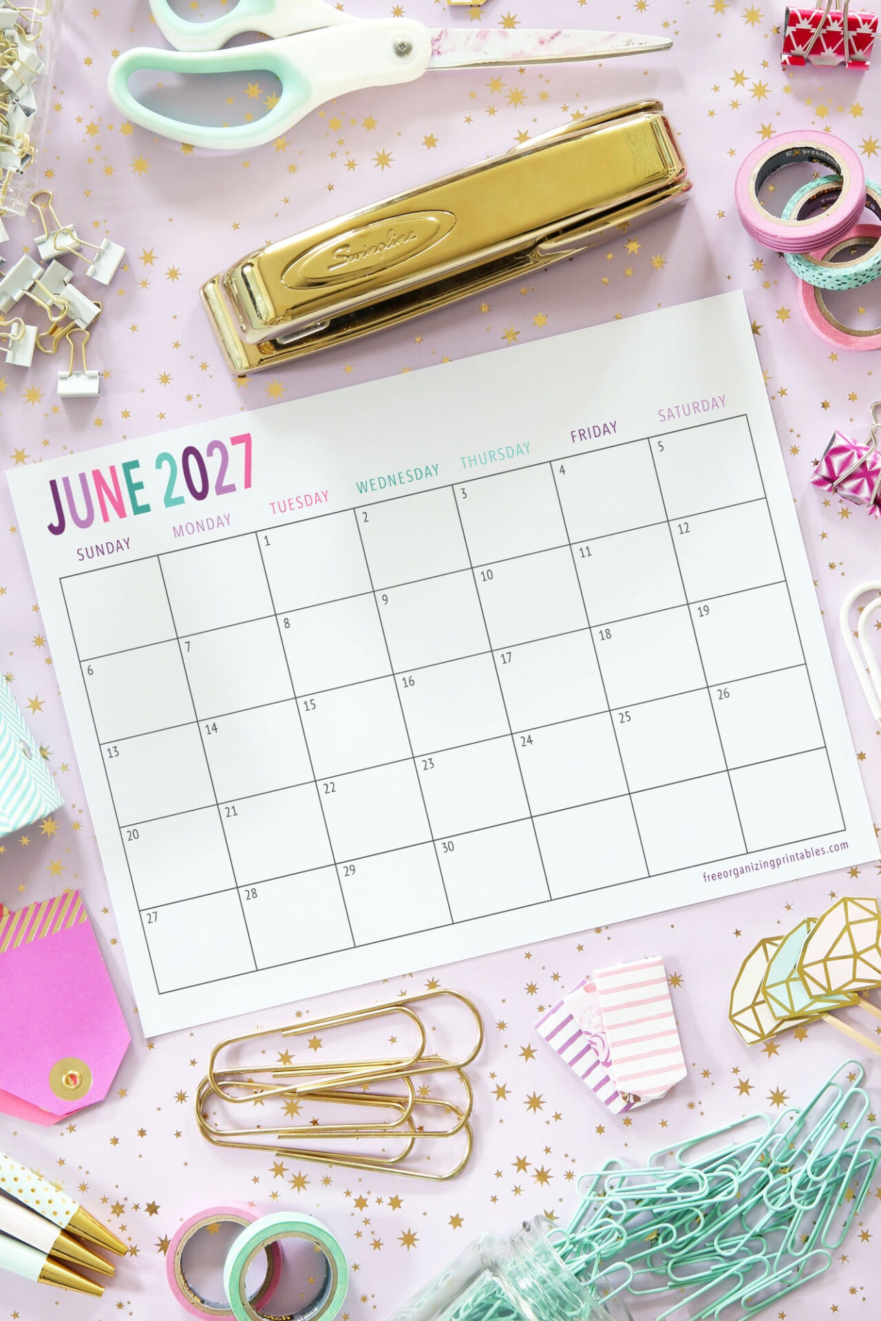 A June 2027 calendar lies on a pink desk scattered with stationery, including scissors, a stapler, binder clips, washi tapes, paper clips, and gold stars.