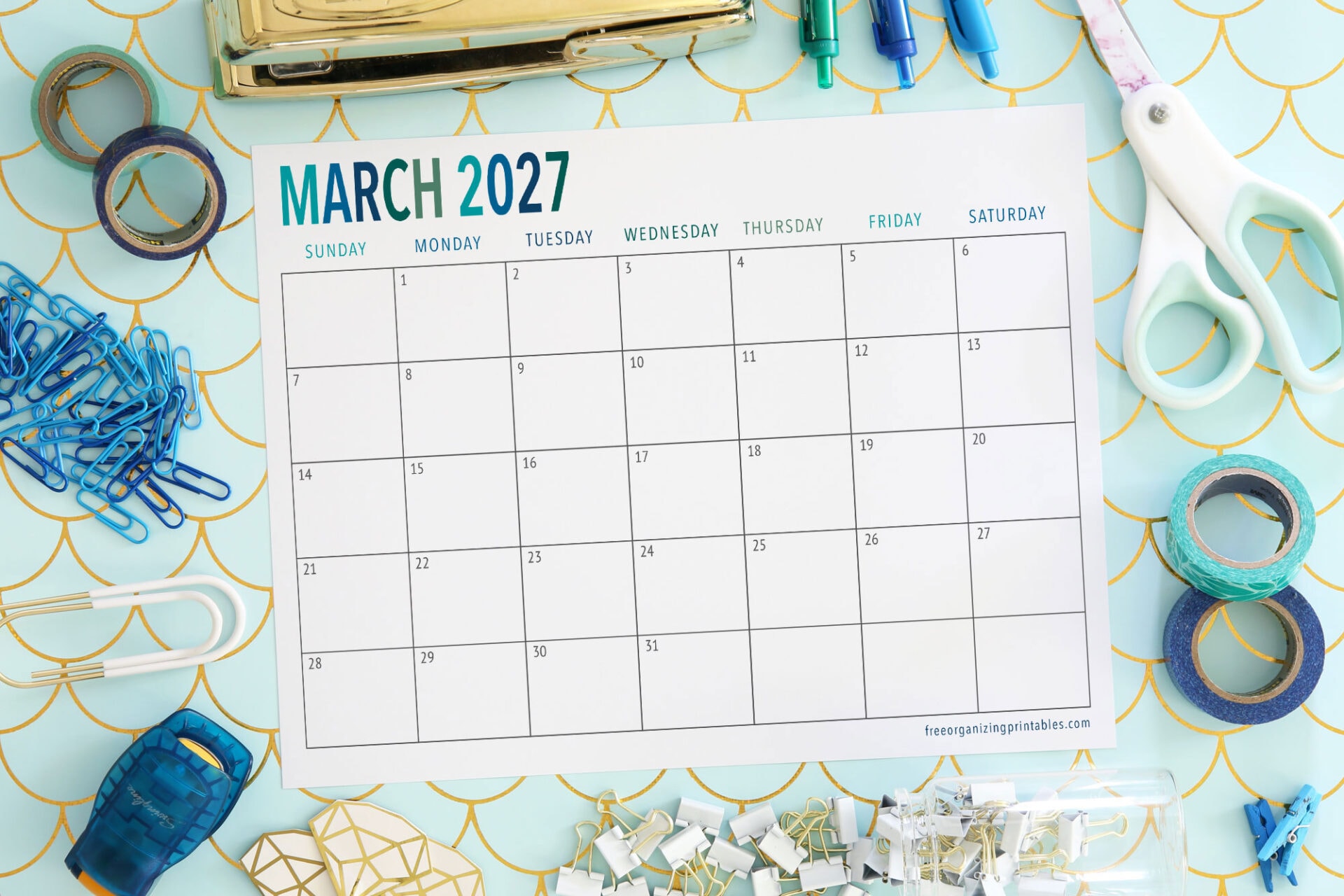A blank March 2027 calendar page is surrounded by office supplies like tape, paper clips, scissors, and a stapler on a blue and gold patterned surface.