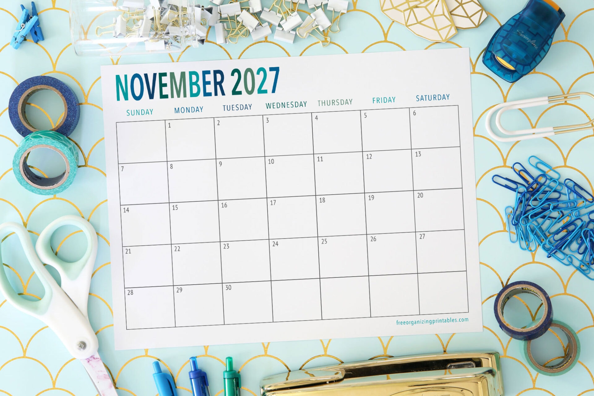 A blank November 2027 calendar page is surrounded by office supplies like scissors, binder clips, pens, washi tape, and paper clips on a light blue patterned background.