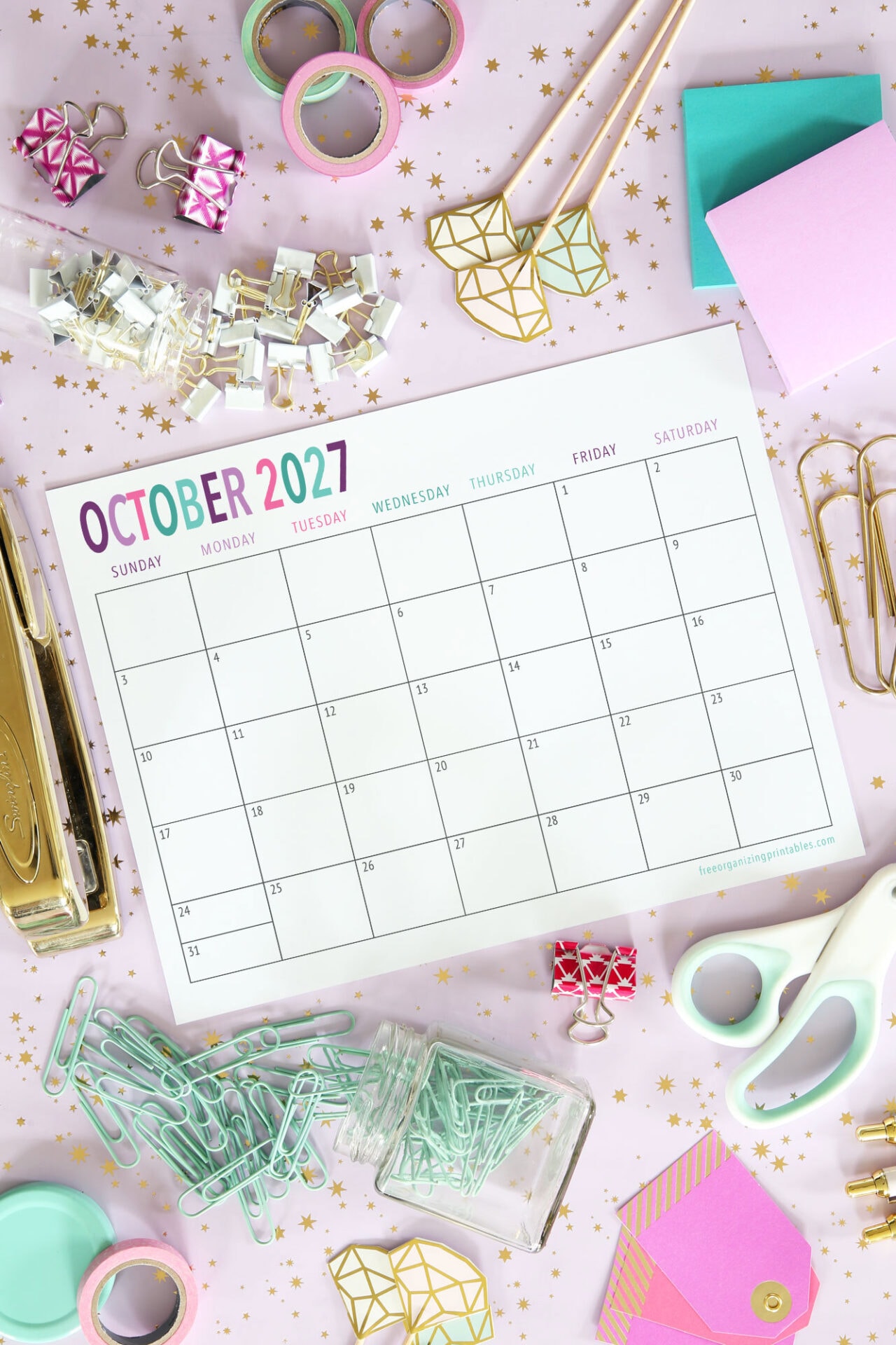 Desktop with a blank October 2027 calendar surrounded by office supplies: paper clips, binder clips, washi tape, sticky notes, scissors, gold stapler, and star confetti on a pink background.