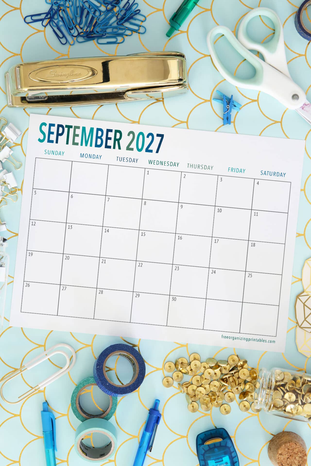A blank September 2027 calendar on a desk surrounded by office supplies like scissors, stapler, paper clips, tacks, and binder clips on a blue patterned background.